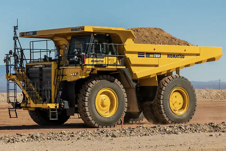 Um grande caminhão fora de estrada amarelo da Komatsu, carregado de terra, segue por uma estrada de terra em uma área de mineração, sob um céu azul claro.
