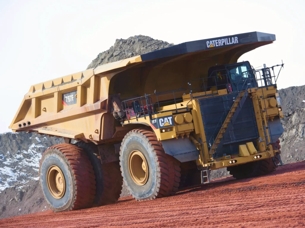 Caminhão de mineração amarelo em operação em um canteiro de obras ao ar livre com céu nublado ao fundo, destaque para máquinas pesadas de mineração.
