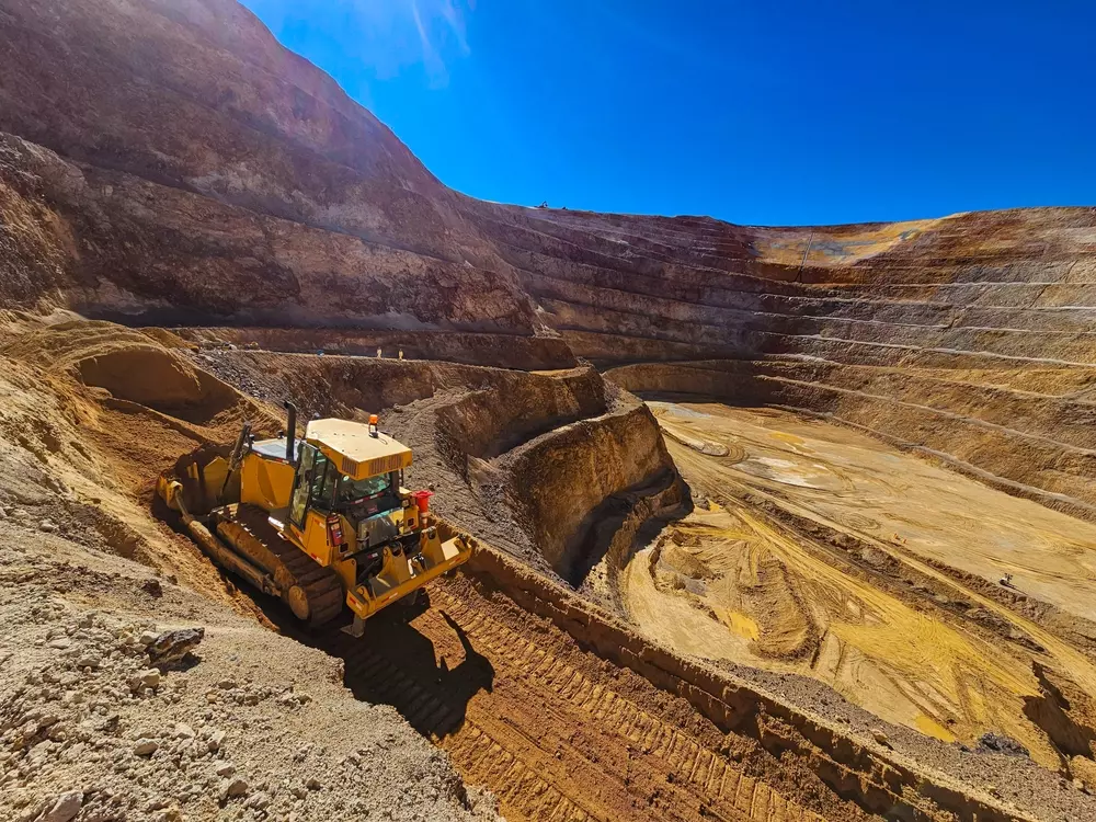 Máquina de escavadeira trabalhando em uma pedreira de grande porte, com camadas de rochas expostas e uma paisagem de céu azul sob o sol intenso.