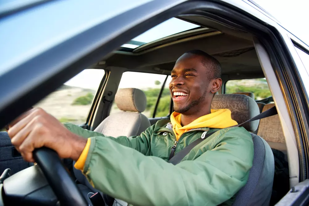 Homem sorridente dirigindo um carro em uma estrada aberta, com expressão de felicidade e liberdade.