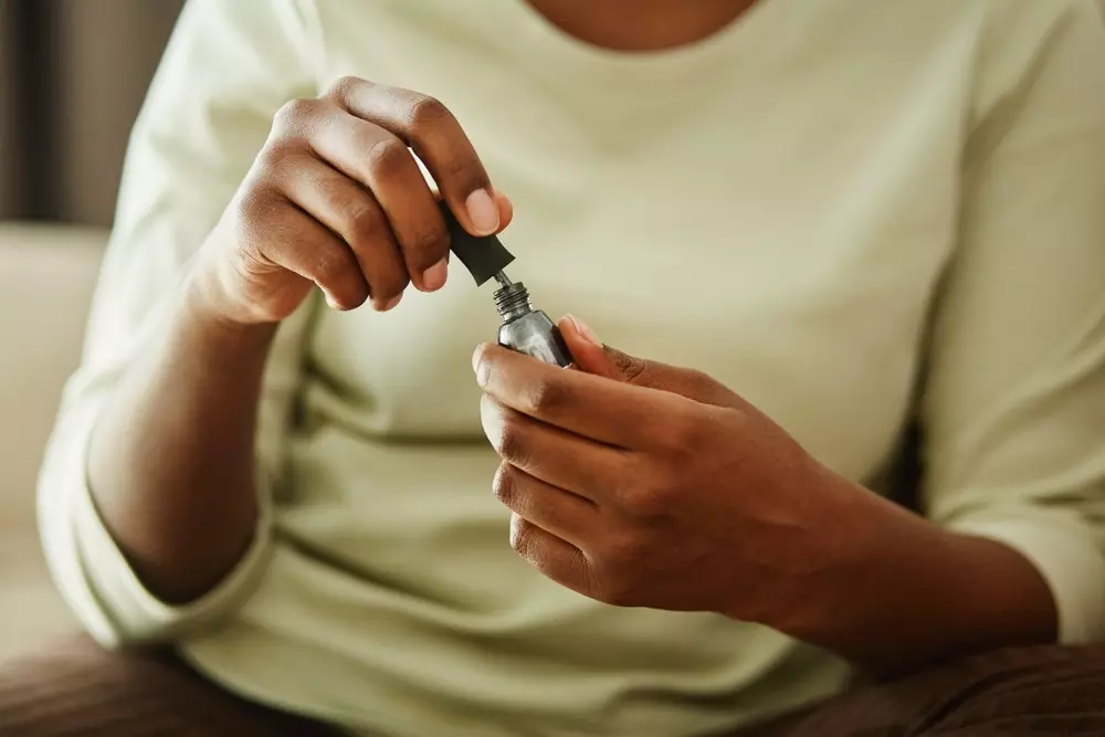 Fotografia em ângulo aproximado de uma jovem negra segurando um frasco de esmalte prateado enquanto faz as unhas em casa.