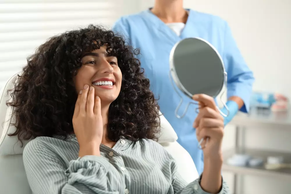 Paciente jovem sorridente com cabelo cacheado observando seu rosto no espelho no consultório odontológico, enquanto profissional de saúde dental está ao fundo.