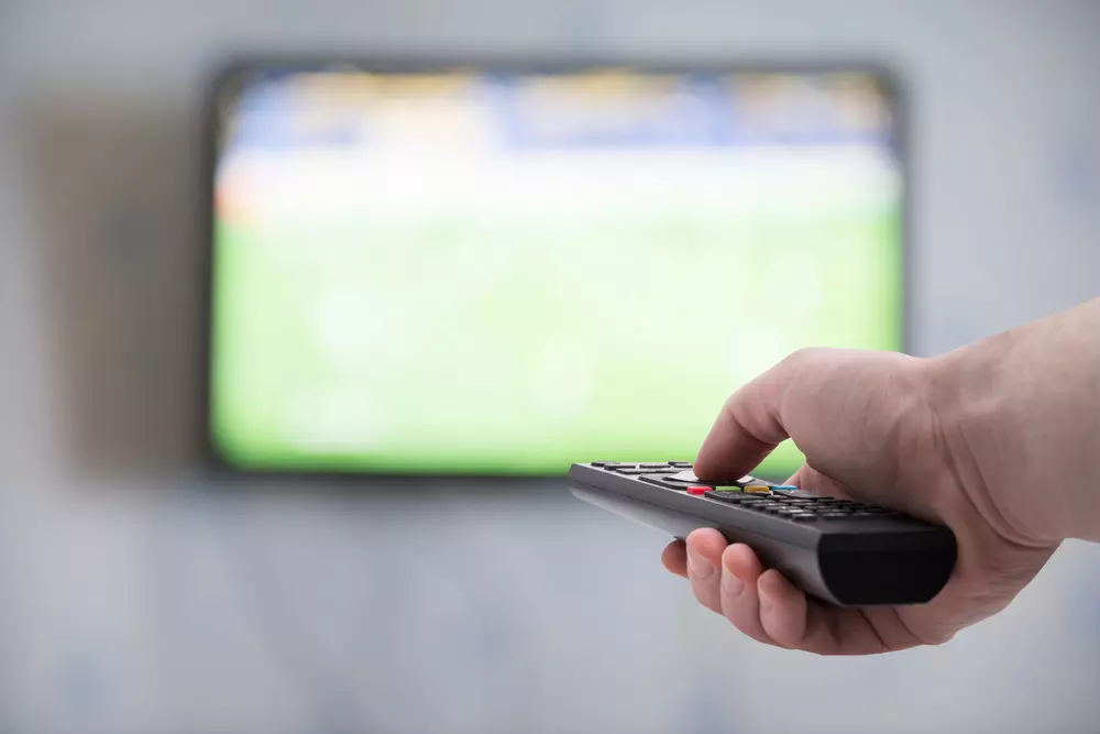 Pessoa assistindo a um jogo de futebol na televisão usando um controle remoto. Imagem focada na mão segurando o controle remoto e na tela da TV ao fundo.a