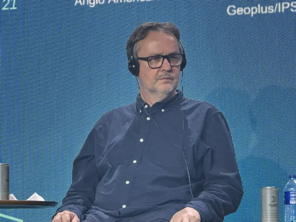 Beto com óculos e fones de ouvido, com expressão séria, participando de um evento ou painel de discussão.