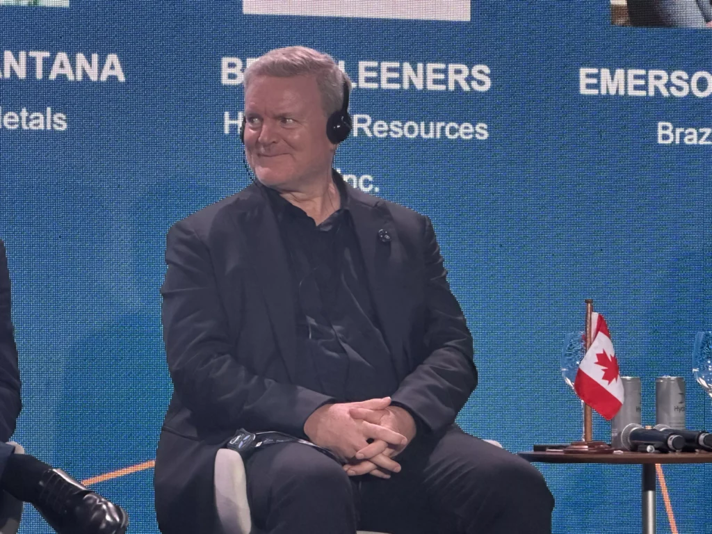 Brian Leeners em evento profissional usando fone de ouvido, sentado em cadeira com fundo azul de painel de conferência, com bandeira do Canadá ao lado.