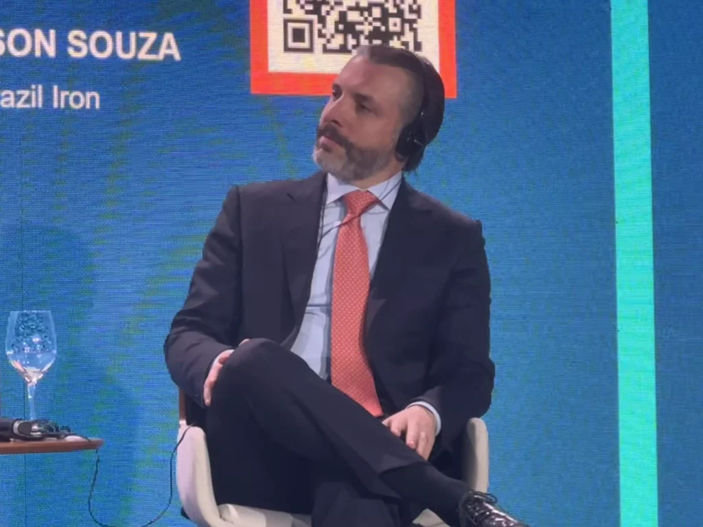 Emerson das Neves Souza em traje formal com cabelo e barba grisalhos, usando fone de ouvido, participando de evento ou debate, com fundo azul e detalhes coloridos.