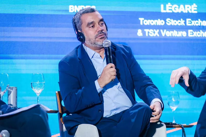 Flávio Moraes da Mota participando de evento, usando terno azul e headset, falando ao microfone em uma conferência.