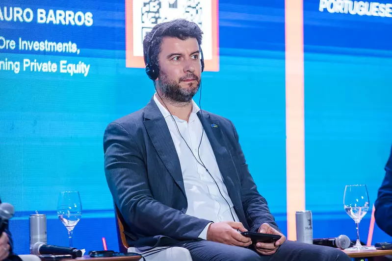 Marco Diogo adulto participando de um painel de discussão em um evento de investimentos, usando blazer e notebook, com fundo azul e painel digital.