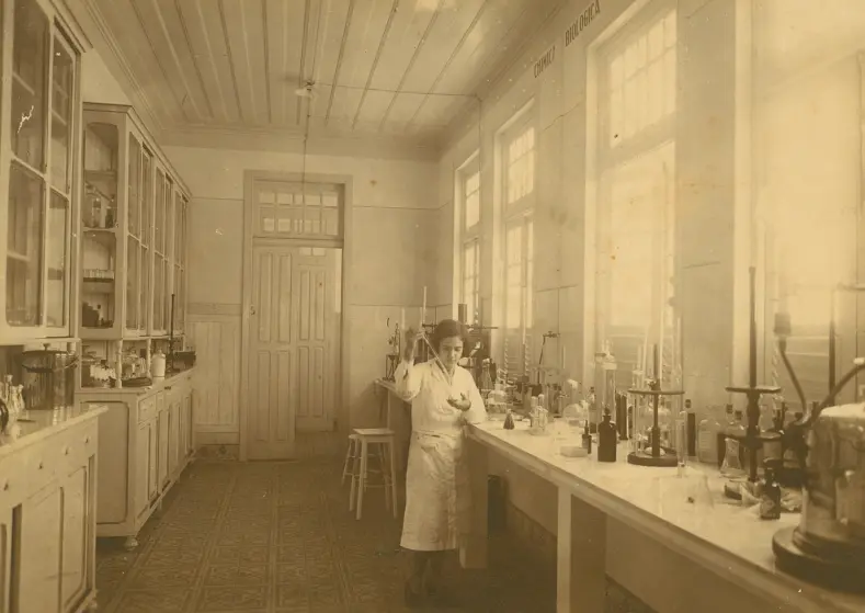 Clara Pandolfo em um laboratório antigo, com bancada cheia de instrumentos científicos e estantes com frascos