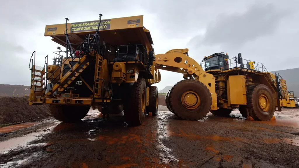 Máquinas pesadas de mineração operando em um projeto de mineração a céu aberto sob céu nublado, destacando a grandeza e a robustez dos equipamentos.