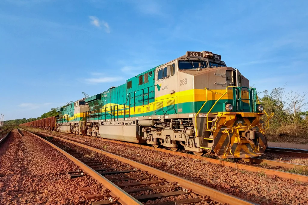 Locomotiva Wabtec na EFVM em operação, destaque para sua pintura verde e amarelo, com trilhos de ferro ao fundo e céu azul ao céu ao fundo.