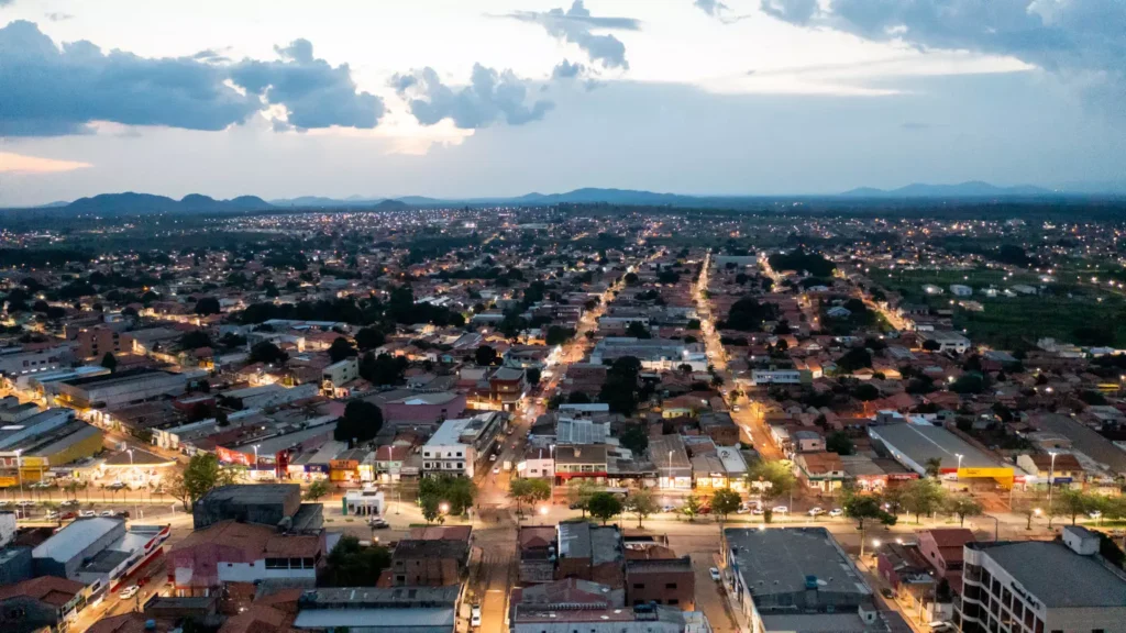 Canaã dos Carajás ao entardecer, mostrando ruas iluminadas e a cidade rodeada por áreas verdes e montanhas ao fundo.