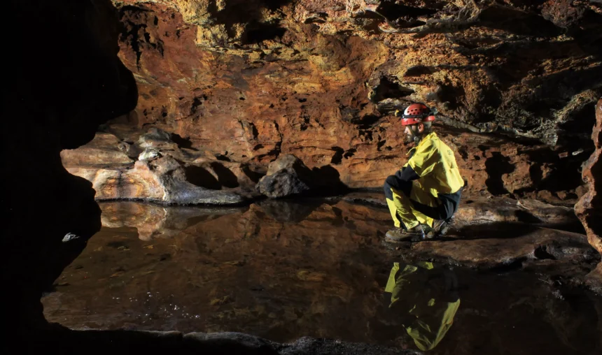 Explorador em uma cavidade subterrânea com paredes rochosas e uma pequena piscina de água, explorando uma caverna de formações rochosas e ambiente úmido.
