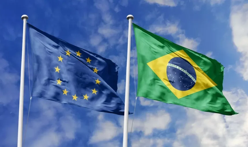 Bandeiras da União Europeia e do Brasil hasteadas lado a lado sob um céu azul com nuvens.
