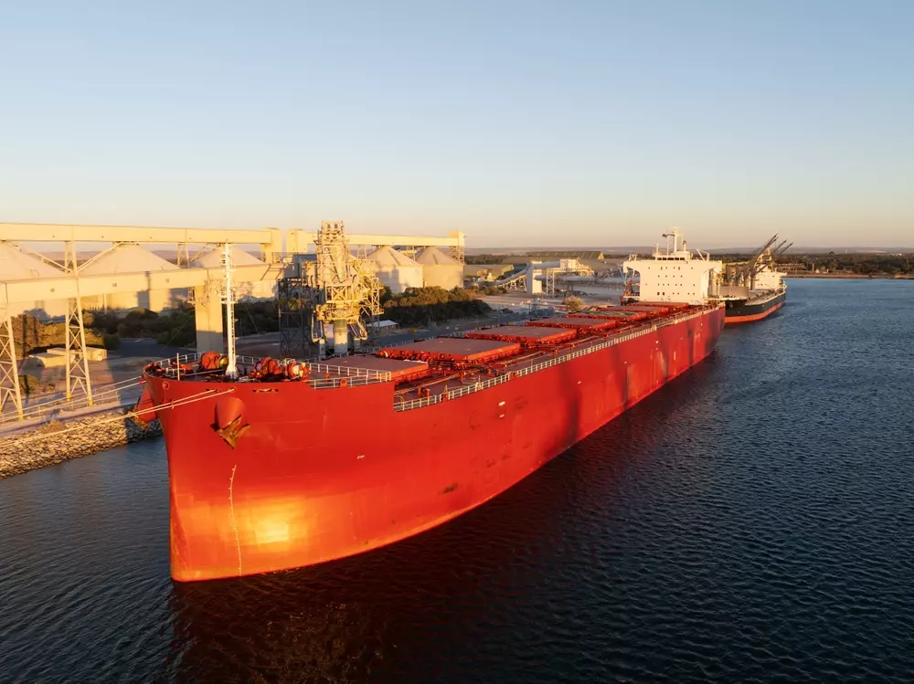 Navio de exportação de minério no porto, carregando carga de minério em uma operação de exportação marítima, com instalações industriais ao fundo.