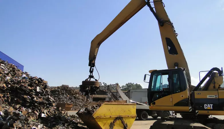 Escavadeira amarela utiliza eletroímã para movimentar sucata metálica em um pátio de reciclagem tupy