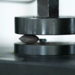 Máquina de compressão testa a resistência de uma pequena pelota escura de minério entre duas placas de metal.