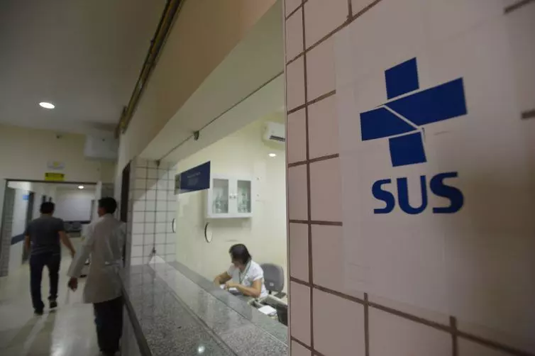 interior de uma unidade do SUS (Sistema Único de Saúde) com profissionais de saúde atendendo pacientes e um logo azul do SUS visível na parede