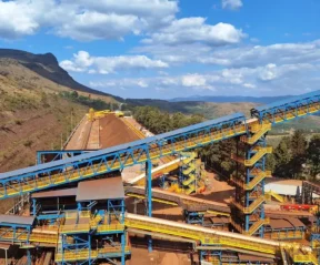 Grande instalação industrial de mineração com correias transportadoras azuis e amarelas transportando terra, cercada por pilhas de solo marrom-avermelhado - capturando a escala das projeções da Vale em meio a árvores, colinas e um céu parcialmente nublado em uma paisagem montanhosa.