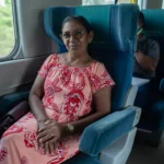 Mulher sorridente sentada em poltrona azul dentro de um ônibus, usando vestido estampado vermelho e branco, com olhar para a câmera durante a viagem.
