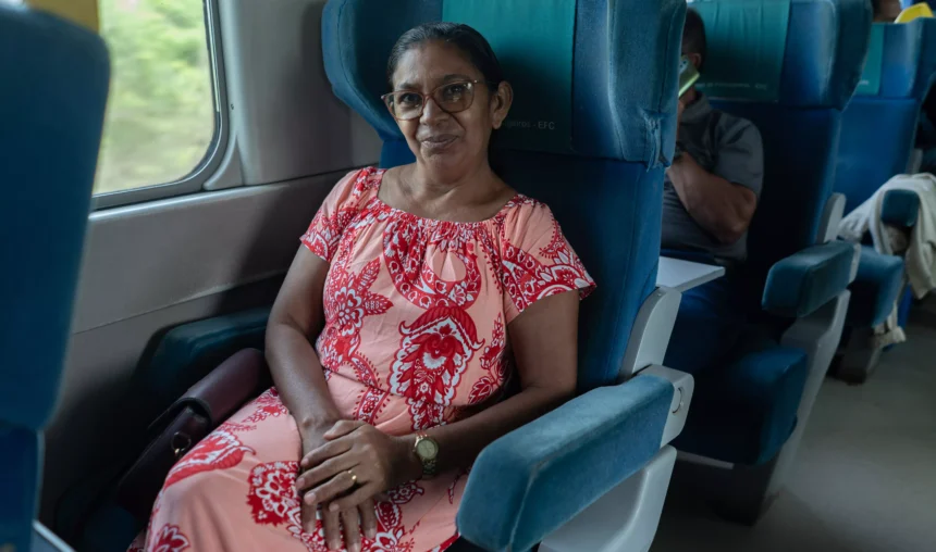 Mulher sorridente sentada em poltrona azul dentro de um ônibus, usando vestido estampado vermelho e branco, com olhar para a câmera durante a viagem.