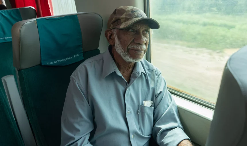 Homem idoso com barba e boné camuflado sentado em um ônibus, usando camisa azul, ao lado da janela com paisagem desfocada ao fundo.