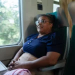 Mulher sentada em um vagão de trem, usando óculos e uniforme escuro, com expressão serena e olhando pela janela. Ao lado há uma poltrona verde