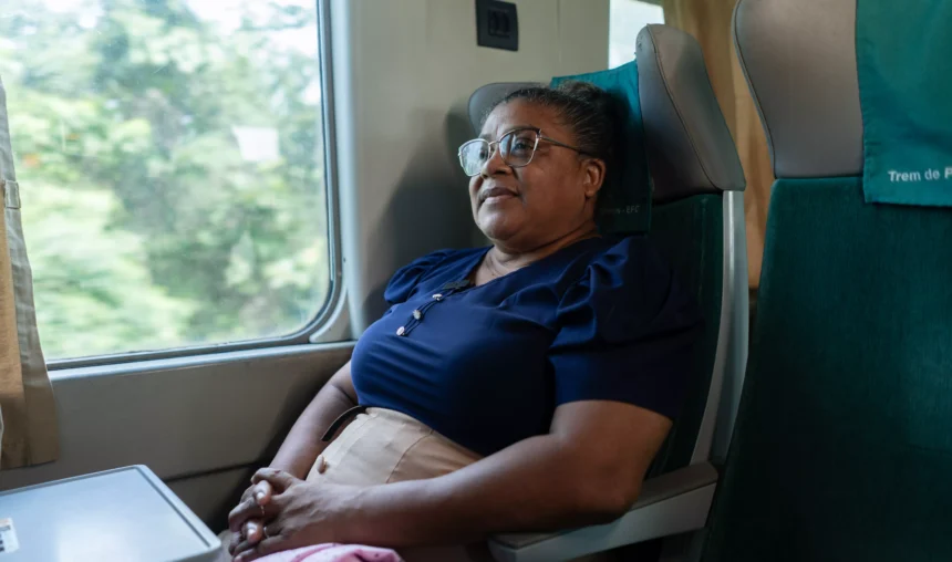 Mulher sentada em um vagão de trem, usando óculos e uniforme escuro, com expressão serena e olhando pela janela. Ao lado há uma poltrona verde