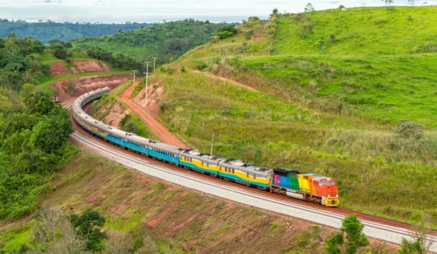 Trem de passageiros colorido atravessando a linha férrea em meio a colinas verdes e áreas de vegetação no interior, visto de cima com céu parcialmente nublado ao fundo.