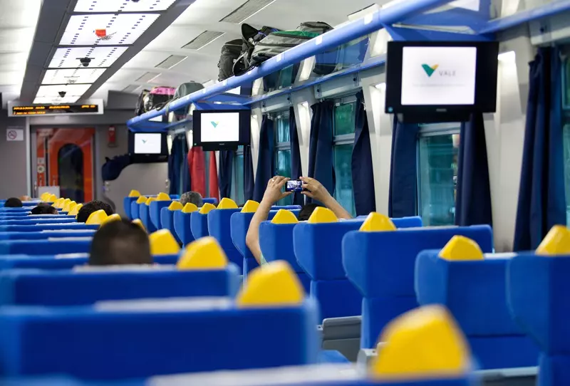 Interior de um trem de passageiros com assentos azuis e amarelos, telas com a indicação da rota e um passageiro tirando foto durante a viagem.