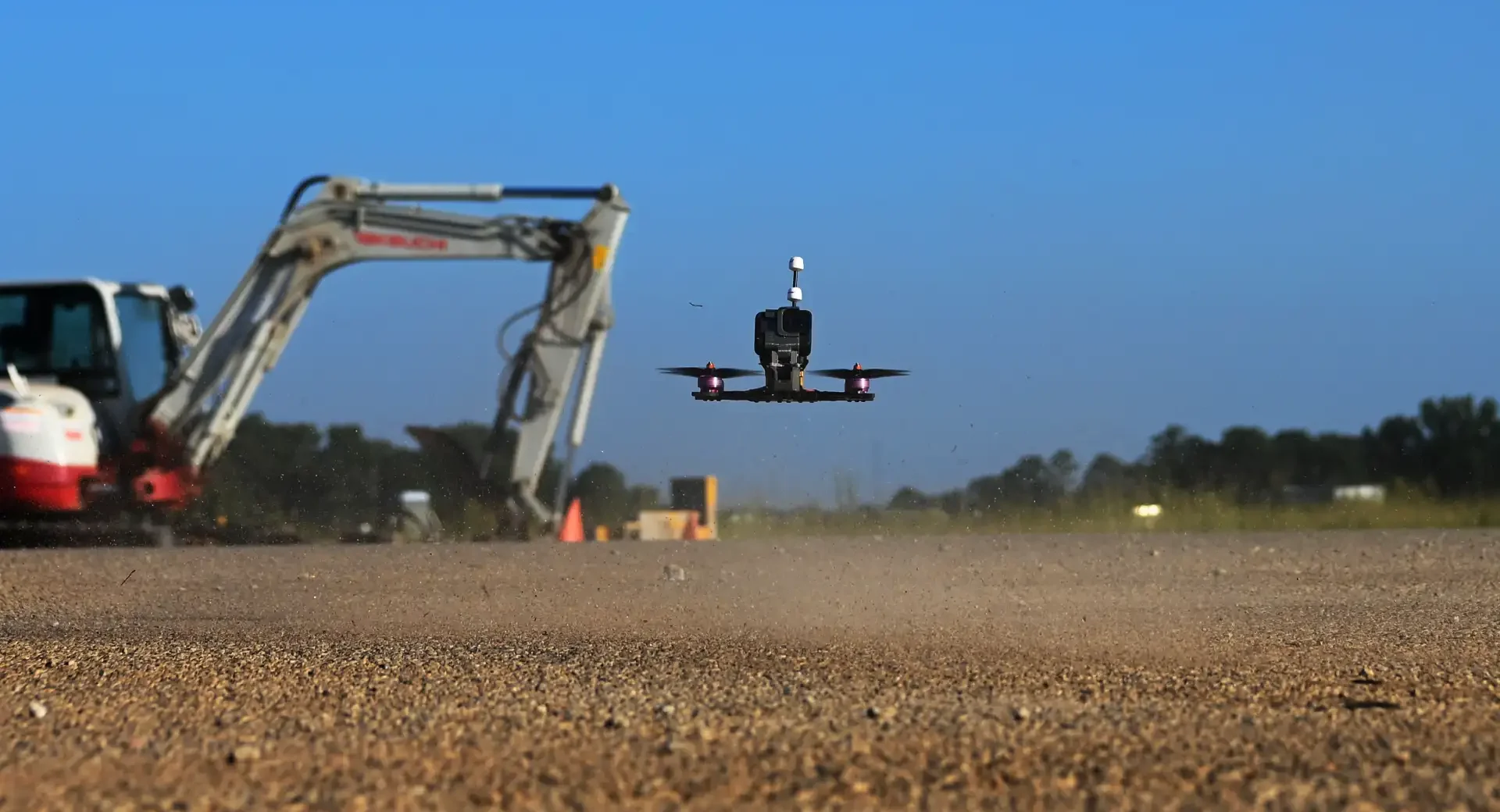 Drone em mineração sobrevoa uma área de trabalho com solo de cascalho ao fundo, enquanto uma máquina pesada opera na obra sob céu azul, destacando monitoramento e inspeção