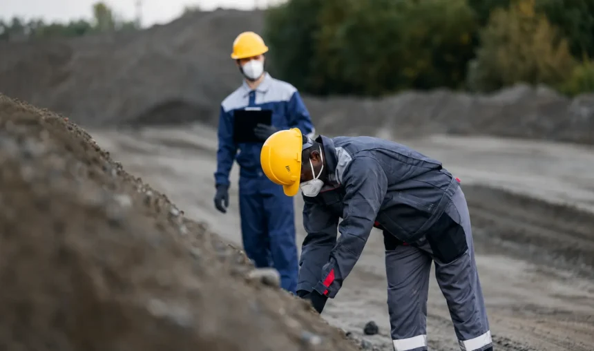 Dois trabalhadores vestindo roupas de proteção, capacetes amarelos e máscaras faciais estão em um canteiro de obras. Um engenheiro de registro examina uma pilha de terra enquanto o outro observa ao fundo. As árvores são visíveis à distância.