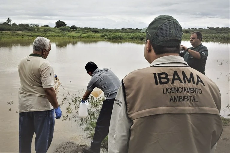 Equipe do IBAMA em campo realiza atividade de licenciamento ambiental e monitoramento junto a um rio, com profissionais usando uniformes do órgão e luvas próximo à vegetação e à margem da água.