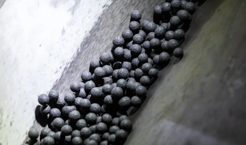 Um close-up de vários pellets pequenos, escuros e redondos de minério de alto teor descendo por uma calha ou esteira industrial inclinada. A superfície metálica áspera capta a luz, criando destaques e sombras impressionantes nos pellets.