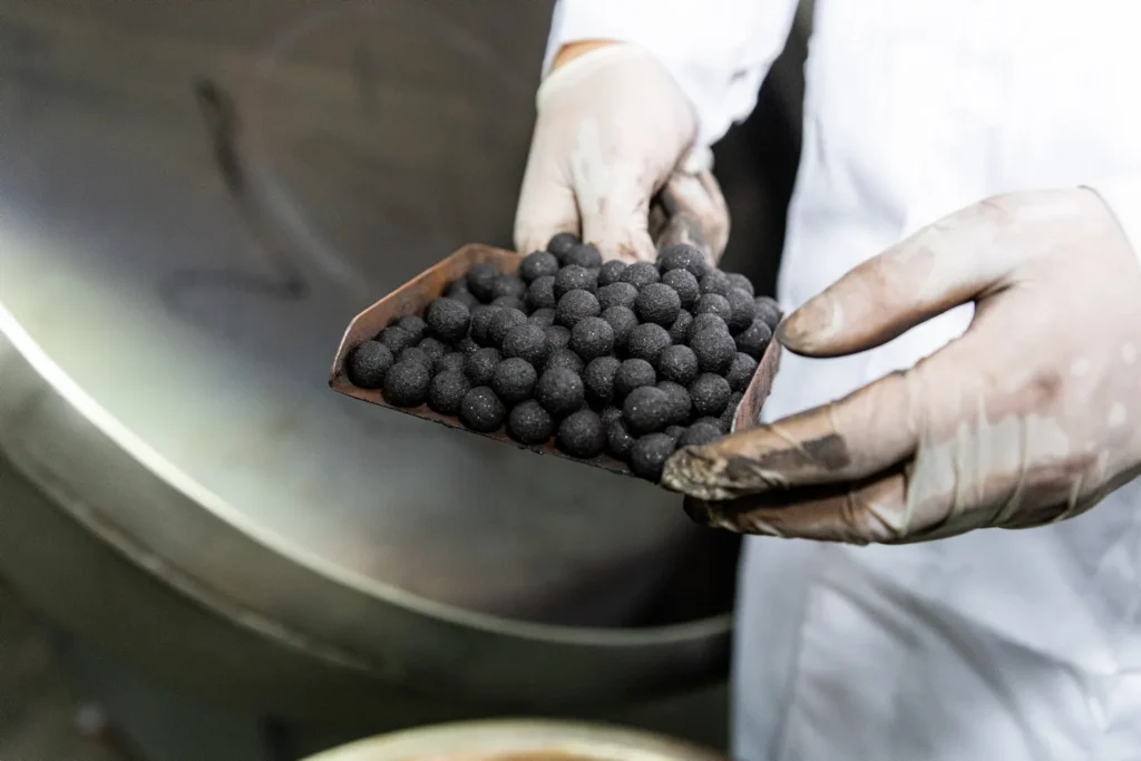 Uma pessoa usando luvas brancas e jaleco branco segura uma bandeja cheia de pellets pequenos, redondos e pretos, ilustrando o processo de pelotização em um ambiente industrial ou de laboratório.