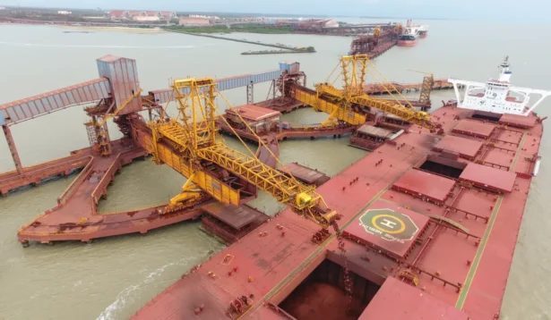 Navio de carga atracado em um porto industrial, com guindastes amarelos descarregando ou carregando materiais no navio em meio a tensões geopolíticas; água e outros navios são visíveis ao fundo.