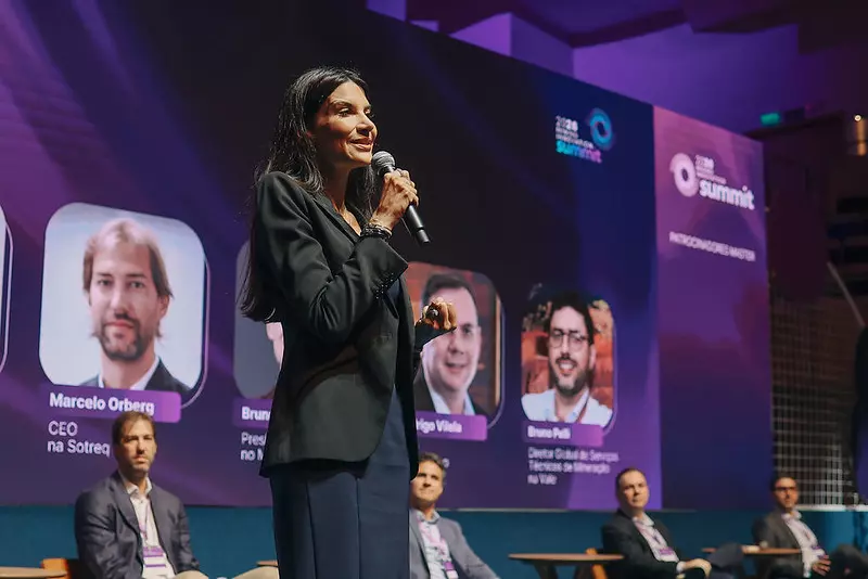 Uma mulher fala em um microfone no palco de uma conferência sobre geoeconomia, com quatro homens sentados atrás dela. Uma tela grande exibe fotos, nomes e títulos dos palestrantes em um pano de fundo roxo.