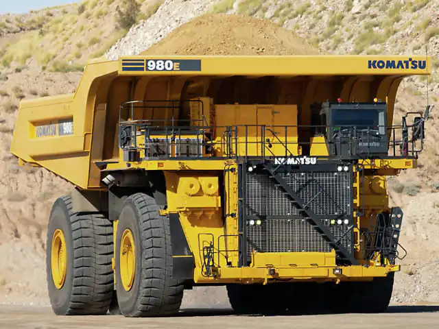 Um grande Komatsu 980E amarelo, um dos principais caminhões fora de estrada para mineração, transporta uma carga cheia de terra por uma estrada de terra em uma mina, tendo ao fundo um terreno rochoso e vegetação esparsa.