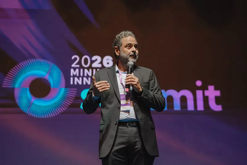 Um homem de terno fala em um microfone no palco de um evento. Atrás dele, luzes coloridas iluminam as palavras 2026 MIN... INN... summit em uma tela grande, destacando os principais temas de geoeconomia e inovação.
