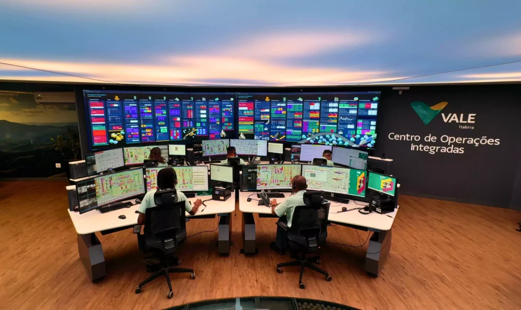 Quatro pessoas sentadas em mesas curvas com vários monitores exibindo dados técnicos em uma moderna sala de controle. Uma grande parede de telas coloridas e uma placa com os dizeres "Vale Centro de Operações Integradas" destacam esse ambiente de modelo usual.