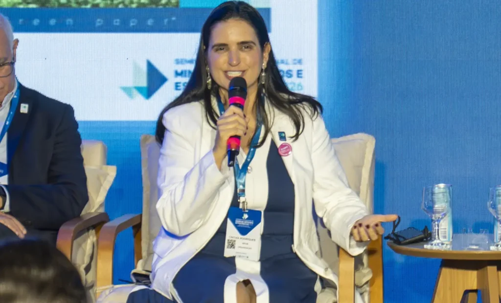 Cinthia de Paiva Rodrigues, gerente de Pesquisa e Desenvolvimento do Ibram, em palestra no palco com microfone e credencial, em evento corporativo com telão ao fundo.