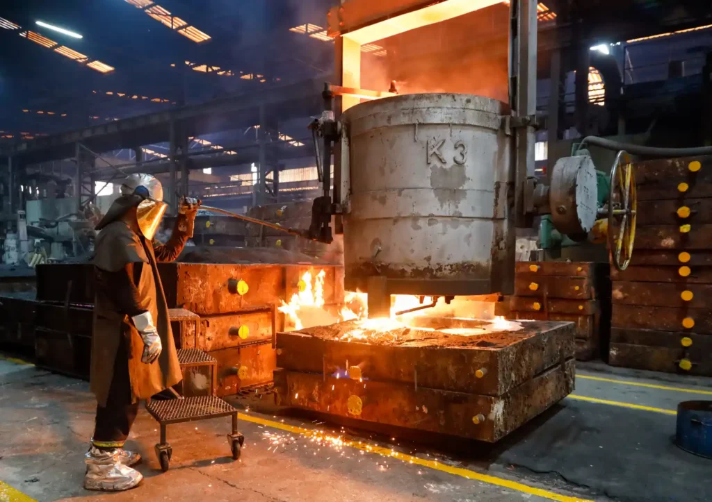 Fundição de aço em processo industrial com trabalhador usando luvas e avental de proteção ao lado de forno com metal incandescente, moldes e equipamento de vazamento na área de fundição