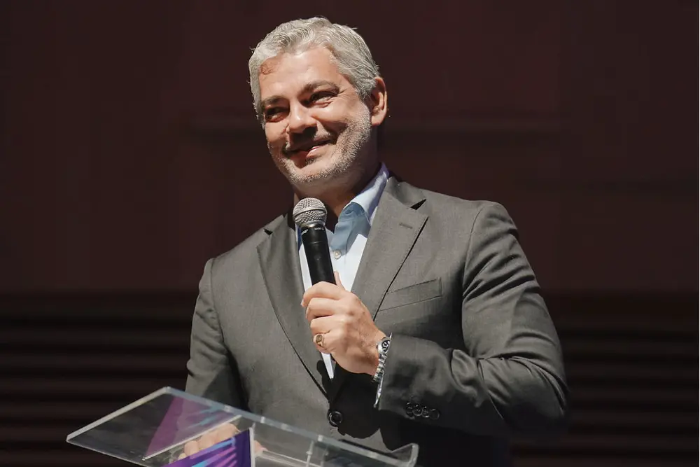 Um homem de cabelos grisalhos e barba, vestindo um terno cinza e camisa azul, sorri enquanto segura um microfone e fala no Mining Innovation Summit de um pódio claro para um público em um ambiente pouco iluminado.