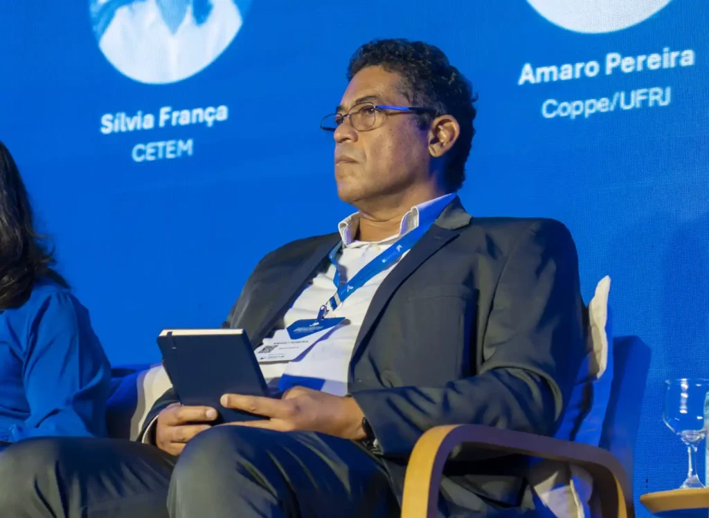 Professor Amaro Pereira, da COPPE/UFRJ, durante apresentação em evento, em painel com crachá e identificação na tela ao fundo, ao lado de participantes.