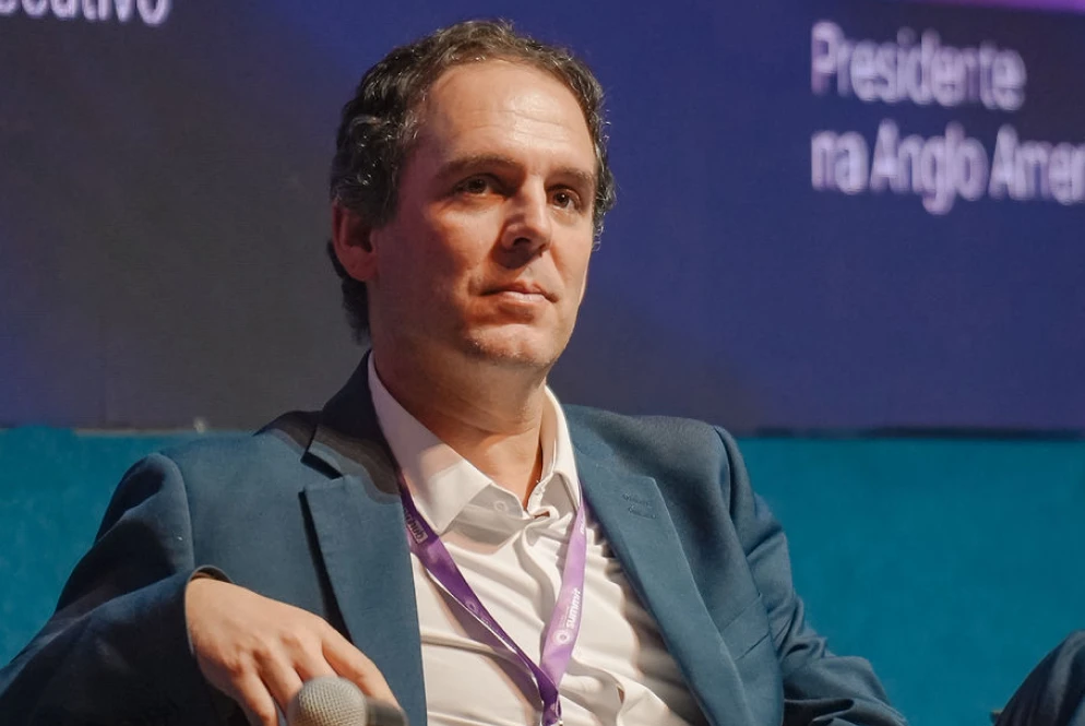 Um homem de terno azul e cordão lavanda senta-se em um palco segurando um microfone, com uma expressão séria enquanto discute geoeconomia. O texto está parcialmente visível em um fundo desfocado.