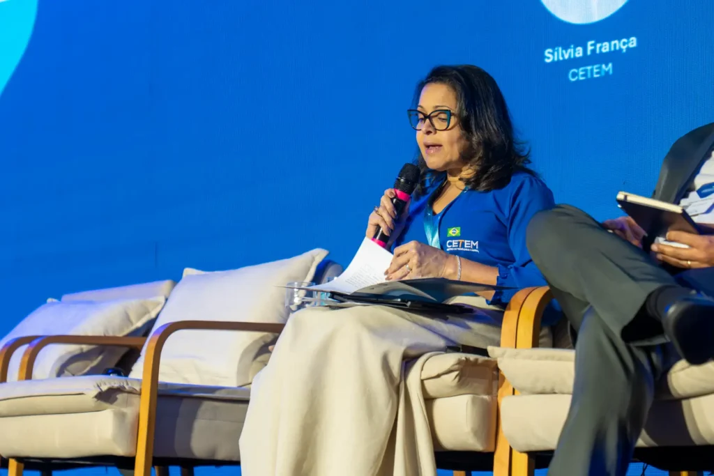 Silvia França, diretora do Cetem, durante palestra em evento, usando microfone e lendo anotações em palco com fundo azul e participantes ao lado.