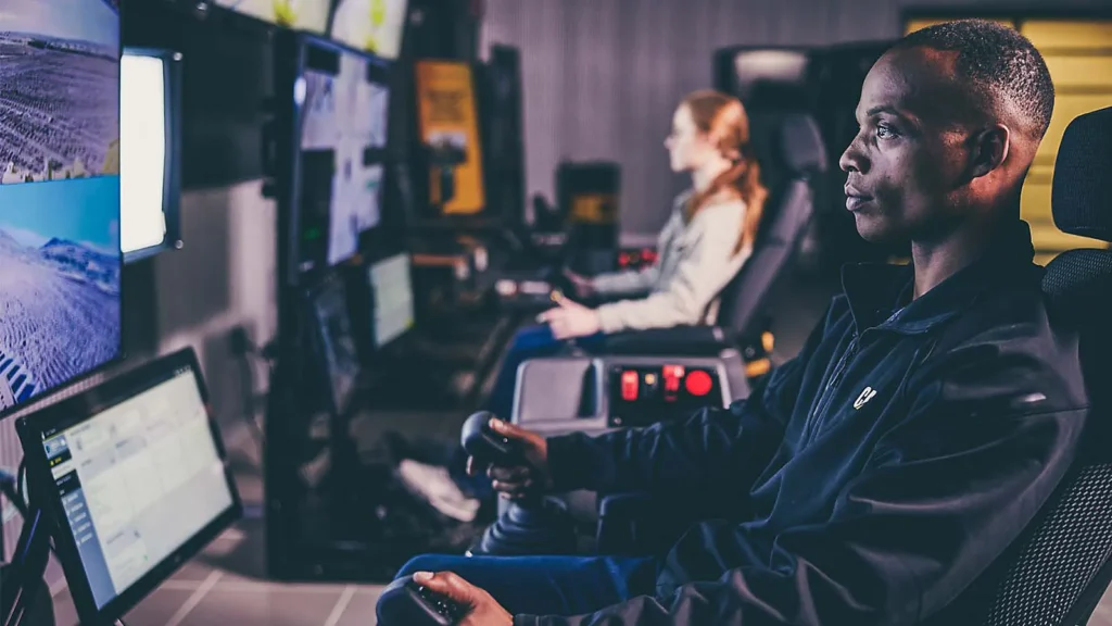 Uma pessoa em primeiro plano opera um joystick em uma estação de trabalho, concentrada em vários monitores, enquanto outra trabalha nas proximidades. O ambiente da sala de controle de alta tecnologia evoca a inovação esperada na Exponor 2026.
