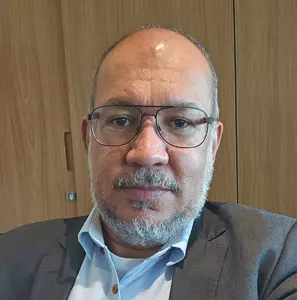Um homem de óculos, barba bem aparada e cabelo curto, vestindo um paletó e uma camisa azul-clara, olha para a câmera. Ele está em um ambiente interno, com painéis de madeira marrom-clara ao fundo, lembrando a presença serena de um imã brasileiro.