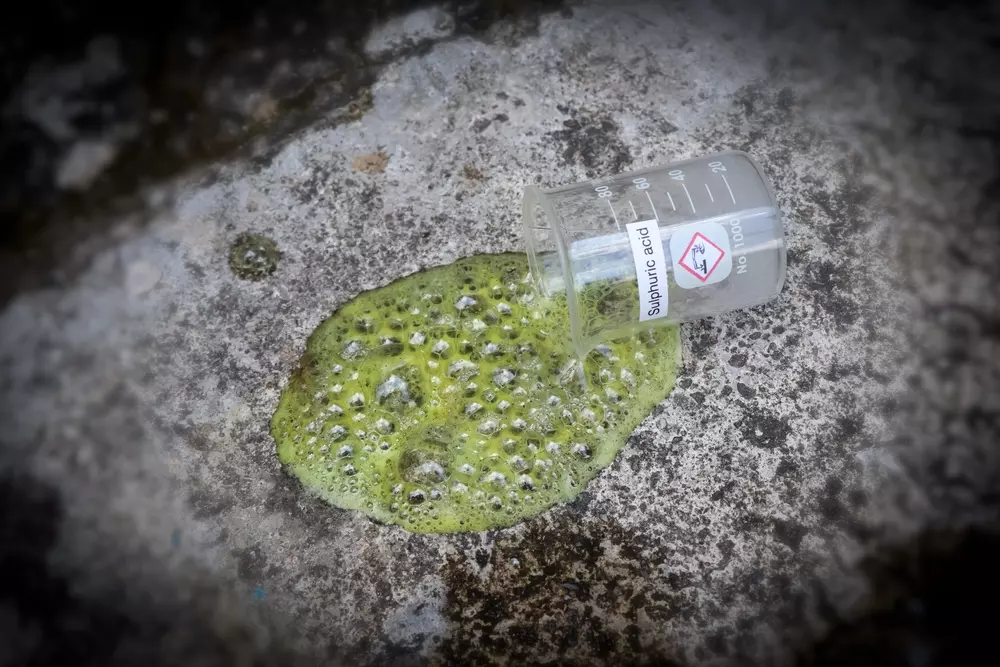 Um copo de precipitação com a etiqueta “Ácido sulfúrico” é derrubado, derramando um líquido verde e borbulhante sobre uma superfície áspera e cinza.