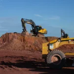 Escavadeiras e equipamentos de construção trabalham em um monte de terra em um canteiro de obras sob um céu azul e limpo, enquanto a equipe avança na extração de terras raras em Serra Verde.